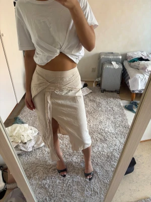 Beige omlottkjol med knyt - Trendig beige omlottkjol med knyt i midjan och slits framtill. Kjolen har en mjuk, något skrynklig struktur och en avslappnad passform som ger en cool och modern look. Perfekt att styla med en enkel t-shirt eller crop top.