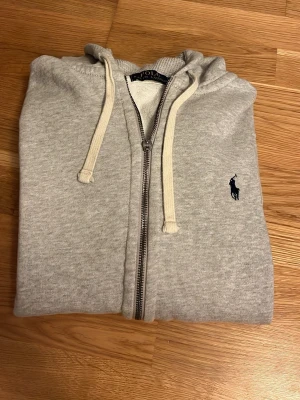 Ralph Lauren hoodie - Snygg grå hoodie med dragkedja från Polo Ralph Lauren. Klassisk design med huva och vita dragsnören samt den ikoniska mörkblå loggan broderad på bröstet. Tillverkad i mjukt bomullsmaterial som känns skönt mot huden.   ‼️OBS‼️dragkedjan har gått sönder (sista bilden) men funkar fortfarande helt utan problem:) pris är diskuterbart!😇