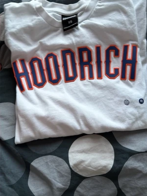 Vit HOODRICH t-shirt med tryck - Säljer en vit t-shirt från HOODRICH med stort tryck i blått och orange över bröstet. T-shirten har klassisk passform och rund hals. Tillverkad i mjuk bomull, perfekt för en avslappnad streetwear-look.