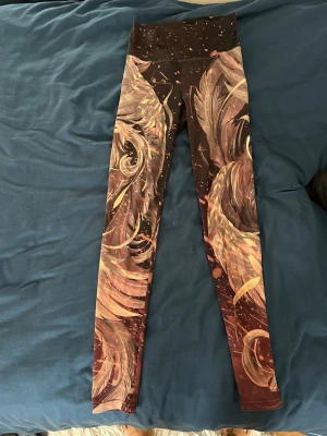 Mönstrade leggings från Nerk Studio - Snygga leggings med hög midja från Nerk Studio. De har ett unikt abstrakt mönster i brunt, beige och guldtoner - innehållande Fågeln Fenix. Tillverkade i stretchigt material som sitter skönt och formar sig efter kroppen. Perfekta för dig som vill sticka ut.