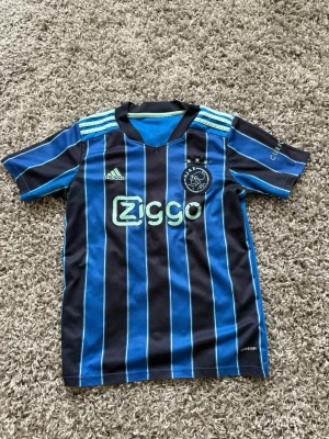 Ajax blå randig matchtröja Adidas - Snygg blå och svart randig matchtröja från Ajax med ljusgröna detaljer och Ziggo-logga på bröstet. Tröjan är i bra skick men sliten på vissa tryck osv