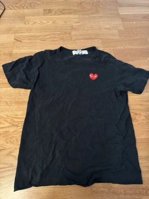 Svart Comme des Garçons PLAY t-shirt - Svart t-shirt från Comme des Garçons PLAY med det ikoniska röda hjärtat med ögon broderat på bröstet. Klassisk passform och rund halsringning. Perfekt för dig som gillar streetwear och stilrena plagg.