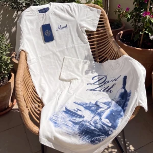 Vit t-shirt med blått tryck - Säljer en vit t-shirt med blått tryck och texten 'Marcel' på bröstet. Baksidan har ett stort motiv med vinflaska, glas och havslandskap i blå toner. Klassisk passform och rund hals. Perfekt för dig som gillar stilrena och unika prints.