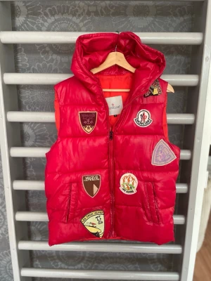 Röd dunväst med patches från Moncler kan användes som l med - Cool röd dunväst från Moncler med huva och dragkedja. Västen är dekorerad med flera färgglada patches och märken både fram och bak, inklusive ett stort gult M på ryggen. Perfekt för dig som vill sticka ut med en sportig och trendig look.