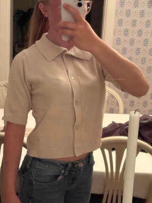 Kofta  - Kortärmad beige kofta från 157 med stora knappar framtill och bred krage. Stickad i mjukt material och har ribbade kanter vid ärmslut och nederkant. Perfekt för lager på lager och enkel att matcha med olika outfits.