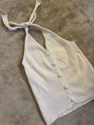 Vit ribbad halterneck topp - Säljer en  vit ribbad halterneck topp med knyt i nacken och dekorativa knappar framtill. Toppen har en djup V-ringning och är gjord i ett stretchigt, mjukt material som sitter tajt och bekvämt. 
