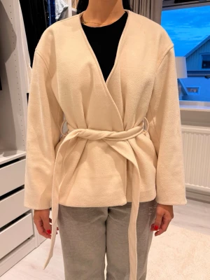 Beige cardiganjacka med knytskärp - Stilren beige cardiganjacka med knytskärp i midjan och öppen front. Jackan har långa ärmar och är tillverkad i ett mjukt, fleece-liknande material. Perfekt för lager på lager och enkel att matcha med olika outfits.