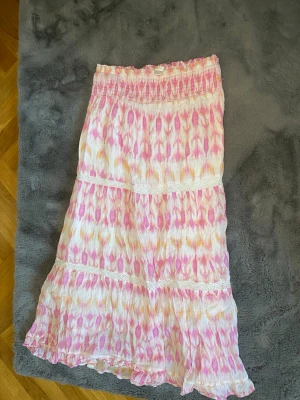 Mönstrad långkjol från Gina Tricot - Säljer en luftig långkjol från Gina Tricot med rosa, gula och vita mönster. Kjolen har resår i midjan, volangdetaljer och är perfekt för varma dagar. Materialet känns mjukt och lätt, vilket gör den skön att bära hela sommaren.