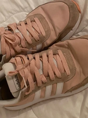Adidas rosa sneakers med mockadetaljer - Säljer ett par snygga Adidas sneakers i ljusrosa med beige mockadetaljer och vita klassiska ränder på sidan. Skorna har rosa snören och vit sula. Perfekta för dig som vill ha en sportig och trendig look. Materialmix av textil och mocka.