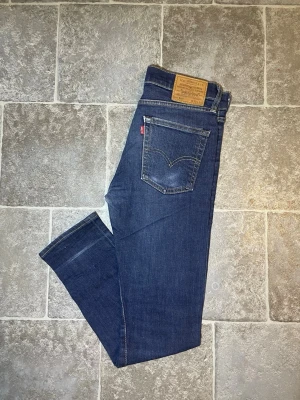 Mörkblå Levi's jeans med rak passform - Snygga mörkblå jeans från Levi's med klassisk rak passform och fem fickor. Jeansen har en ikonisk läderpatch bak i midjan och kontrastsömmar. Tillverkade i slitstarkt denim med en liten stretch för extra komfort. Perfekta för en avslappnad och trendig stil.