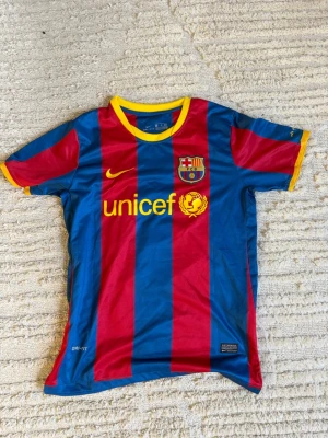 Barcelona Messi 10 - Säljer en klassisk FC Barcelona matchtröja från Nike med Messi och nummer 10 på ryggen. Tröjan har blå och röda vertikala ränder, gula detaljer vid krage och ärmslut samt klubbmärke och Unicef-logga på bröstet. Perfekt för dig som älskar fotboll och Messi.