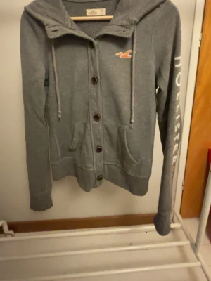 Grå hoodie med knappar från Hollister - Snygg grå hoodie från Hollister med knappar framtill, huva med dragsko och fickor. Hollister-logga på bröstet och text på ena ärmen. Perfekt för en avslappnad och chill stil. Mjuk bomullsblandning som känns skön mot huden. Står storlek m men sitter som xs. Skriv vid minsta fundering