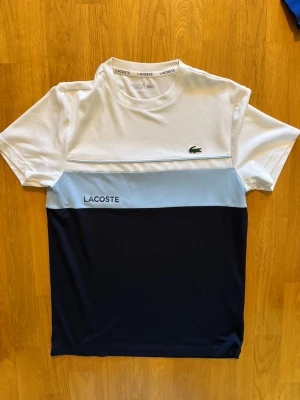 Flerfärgad t-shirt från Lacoste - Snygg t-shirt från Lacoste med färgblock i vitt, ljusblått och mörkblått. Klassisk krokodil-logga på bröstet och Lacoste-tryck på framsidan. Perfekt för en stilren och sportig look.