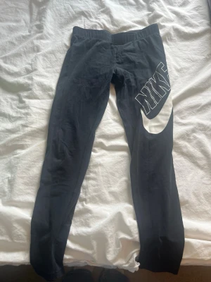 Svarta leggings från Nike - Snygga svarta leggings från Nike med stor vit logga och swoosh på ena benet. De har en tight passform och bred resår i midjan för extra komfort. Perfekta till träning eller chill. Materialet känns mjukt och stretchigt. Använda flera gånger men säljer eftersom dom blivit för korta men jätte fina på och i jätte fint skick.