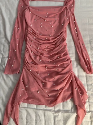 Rosa kort klänning med pärlor - Supersöt rosa kortklänning med långa ärmar och dekorativa pärlor över hela tyget. Klänningen har draperad front, fyrkantig urringning och asymmetrisk nederdel som ger en unik look. Tillverkad i ett tunt, stretchigt material som känns bekvämt.