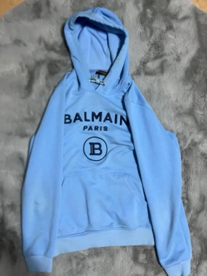 Ljusblå Balmain hoodie med huva - Snygg ljusblå hoodie från Balmain med stor logga och texten 'BALMAIN PARIS' på bröstet. Klassisk känguruficka framtill och ribbade muddar. Huva med dragsko för extra stilpoäng. Perfekt för dig som vill sticka ut med en trendig streetwear-look.