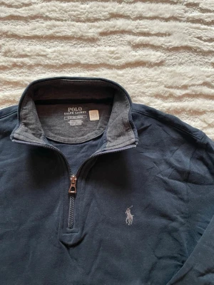 Mörkblå half-zip tröja Polo Ralph Lauren - Mörkblå tröja från Polo Ralph Lauren med halv dragkedja och klassisk broderad logga på bröstet. Tröjan har hög krage med kontrastfärgad insida och är tillverkad i mjukt bomullsmaterial. Perfekt för en avslappnad men stilren look. Inga skavanker. Längd: 65cm. Bredd: 49cm