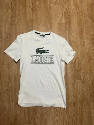 Vit Lacoste t-shirt med logga - Snygg vit t-shirt från Lacoste med klassisk grön krokodil och stor logga på bröstet. Det står storlek xxs fast sitter perfekt för storlek S. T-shirten har rund hals och korta ärmar, tillverkad i mjuk bomull för skön känsla. Perfekt till jeans eller shorts för en clean look. Köpt från Spanien för 850kr