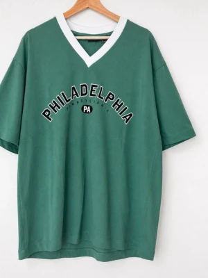Ny oanvänd Grön t-shirt med v-ringning - Grön t-shirt med vit v-ringad krage och texten 'PHILADELPHIA PENNSYLVANIA PA' tryckt på bröstet. T-shirten har en avslappnad passform och korta ärmar, perfekt för en chill och sportig look.