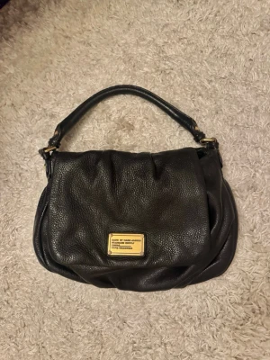 Svart handväska från Marc by Marc Jacobs - Snygg svart handväska i mjukt läder från Marc by Marc Jacobs. Väskan har ett guldfärgat metallmärke framtill och ett kort handtag. Insidan är fodrad med ett coolt svartvitt mönster med bokstäver. Perfekt för dig som gillar stilrena accessoarer. Finns även långt band 🙌🏼