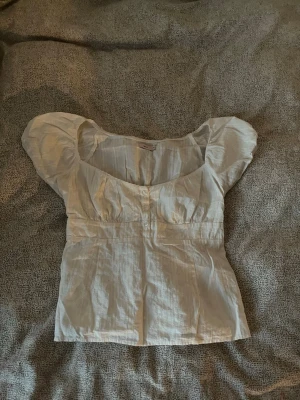 Vit blus med puffärm och knappar - Söt vit blus med puffiga korta ärmar och knappar framtill. Blusen har en lätt v-ringad urringning och är sydd i ett tunt, luftigt tyg som passar perfekt till varma dagar. jättefin men tyvärr för stor för mig 🤍