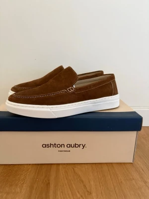 Bruna mocka loafers från Ashton Aubry - Snygga bruna loafers i mocka från Ashton Aubry med vit sula och dekorativa sömmar. Skorna har en klassisk slip-on design och passar perfekt till en avslappnad men stilren look.