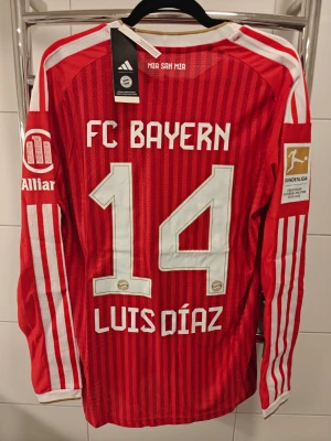 Nya Bayern Munch hemma tröja, spelar version, L - Säljer en röd långärmad FC Bayern München matchtröja från Adidas med vita detaljer och klubbmärke på bröstet. Tröjan har nummer 14 och namnet Luis Díaz på ryggen, samt Bundesliga- och sponsorloggor på ärmarna. Tillverkad i lätt och ventilerande material. 