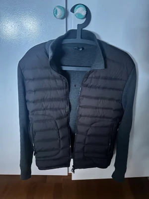 Monclr Cardigan - Litet hål på ryggen av jackan men går att fixa billigt och snabbt. Snygg grå pufferjacka med quiltad front och stickade ärmar samt rygg. Jackan har dragkedja framtill och hög krage för extra värme. Perfekt för dig som vill ha en modern och bekväm look till vardags.