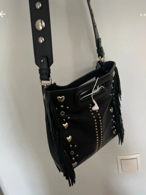 Maje väska - Cool svart axelväska i skinnimitation med guldiga hjärtformade nitar, små runda nitar och långa fransar på sidorna. Väskan har bred axelrem med stora metallknappar och ett handtag upptill. Perfekt för dig som gillar edgy detaljer och unik stil.