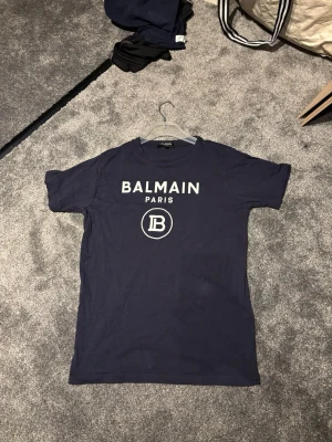 Mörkblå Balmain Paris t-shirt - Snygg mörkblå t-shirt från Balmain Paris med vit logga och text på bröstet. Klassisk passform och rund halsringning. Perfekt för dig som vill ha en stilren och trendig look.