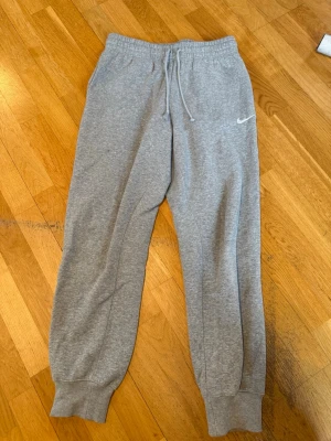 Gråa mjukisbyxor från Nike - Säljer ett par klassiska gråa mjukisbyxor från Nike med vit logga på benet. Byxorna har dragsko i midjan. Tillverkade i mjukt sweatshirtmaterial som är super skönt! Säljer då jag aldrig använder dessa. Tyvärr finns lappen med storlek ej kvar men de passar på mig som är 162 cm! 