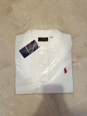 Vit pikétröja från Polo Ralph Lauren - Klassisk vit pikétröja från Polo Ralph Lauren med röd broderad logga på bröstet. Tröjan har krage, knappar framtill och är tillverkad i bomull. Perfekt för en stilren och tidlös look.