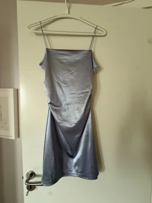 Blå/Silvrig kort klänning  - Säljer en blå/silvrig kort klänning med smala axelband och slät, glansig yta. Klänningen har en enkel och stilren design, perfekt för dig som gillar minimalistiskt mode. 