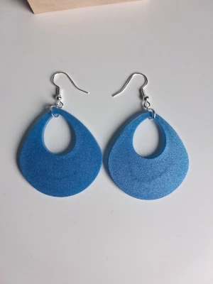 Stora blå droppformade örhängen skimmer blå resin - Säljer ett par stora, droppformade örhängen gjorda i resin/epoxy i skimmer blå färg. Hänget är ca 4.5cm stort. Öronkroken är nickelsäker Perfekta för dig som vill ha ett statement-smycke som sticker ut och ger färg till din outfit.
