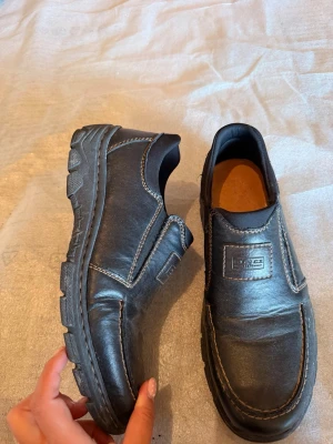 Skor från Rieker - Säljer ett par svarta skor från Rieker i skinn med grov sula och kontrastsömmar. Skorna har slip-on-modell och en bekväm innersula.