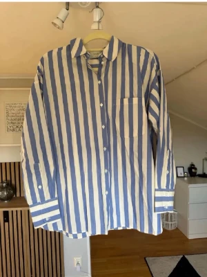 Blåvit randig skjorta från Adpt - Snygg skjorta från Adpt med breda blå och vita vertikala ränder. Skjortan har klassisk krage, lång ärm med manschett och en bröstficka. Tillverkad i ett lätt och bekvämt bomullstyg, perfekt för en avslappnad men trendig look.