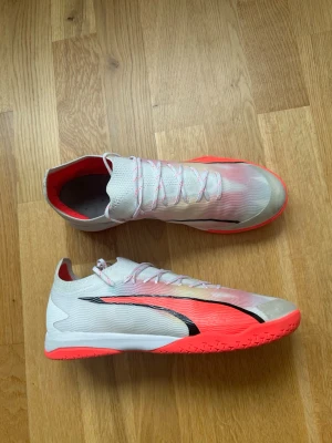 Vita och rosa fotbollsskor med svart detalj - Snygga fotbollsskor i vitt med rosa och neonröda detaljer samt en svart vågig linje på sidan. Skorna har snörning och en låg profil, perfekt för snabba rörelser på planen. Yttermaterialet är syntetiskt och sulan är i gummi för bra grepp.
