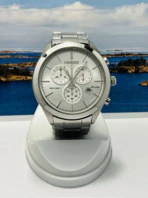 Silverfärgad kronografklocka Champion - Stilren kronografklocka från Champion i silverfärgat metall med länkarmband. Urtavlan är vit med tre mindre subdials, datumvisning och tydliga markeringar. Klockan har en klassisk rund form och passar perfekt för dig som gillar en clean och modern look. Små obentliga repor finns men påverkar ej funktion eller helhetssyn. Nytt batteri, säljes i befintligt skick enligt bilderna! 