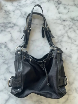 Svart handväska med crocopräglade detaljer - Snygg svart handväska med crocopräglade detaljer och silverfärgade spännen. Väskan har justerbara axelband och dragskostängning. Materialet är en mix av textil och konstläder, vilket ger en cool kontrast. Perfekt för dig som gillar en edgy och trendig look.