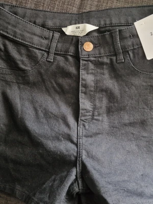 Två par jeansshorts från H&M - Två par klassiska jeansshorts från H&M, ett par i svart denim och ett par i ljusblå denim. Båda har normal passform, fem fickor, bälteshällor och knappstängning framtill. Perfekta för sommaren och lätta att styla med olika toppar.