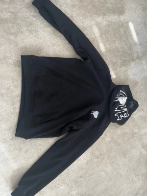 Svart hoodie med tryck och huva - Säljer en svart hoodie med stor ficka framtill och huva. På huvan finns ett vitt tryck med text och ansikte, samt ett litet tryck på bröstet. Ärmarna har ribbade muddar och ett diskret tryck på ena ärmen. Perfekt för en avslappnad streetwear-look.