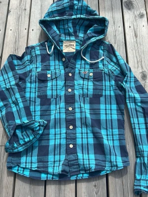 Blå rutig skjorta med huva Hollister - Säljer en blå och turkosrutig skjorta från Hollister med huva och vita knappar. Skjortan har två bröstfickor med knappstängning och dragsko i huvan. Perfekt för en avslappnad och cool stil.