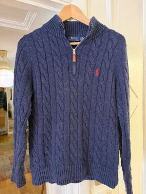 Marinblå kabelstickad tröja Polo - Kabelstickad marinblå tröja från Polo Ralph Lauren med halv dragkedja och hög krage. Tröjan har ett rött broderat logomärke på bröstet, ribbade muddar och nederkant. Perfekt för lager på lager och klassisk stil. Aningen nopprig