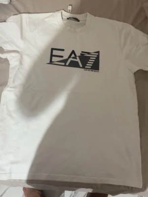 Vit EA7 t-shirt med tryck - Snygg vit t-shirt från Emporio Armani EA7 med stort svart tryck på bröstet. Klassisk rund hals och korta ärmar. Perfekt för dig som gillar stilrena plagg med en sportig touch.