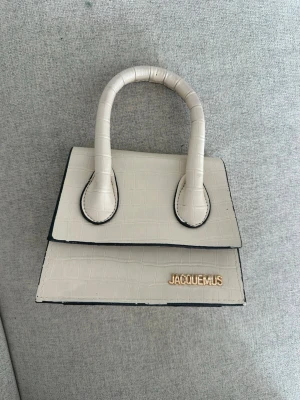Beige Jacquemus handväska i croc-mönster - Stilren beige handväska från Jacquemus med croc-embossad yta och guldfärgad logga framtill. Väskan har en unik, strukturerad form med handtag och svart kantdetalj. Perfekt för dig som gillar trendiga accessoarer med lyxig känsla. (inte äkta)