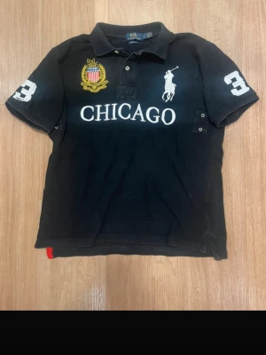 Svart pikétröja med CHICAGO-tryck - Svart pikétröja med vit text 'CHICAGO' på bröstet, broderad gul och röd patch samt stor vit Polo-logga. Nummer 3 i vitt på ena ärmen och detaljer i vitt. Klassisk krage och knappar framtill.