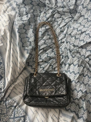 Svart quiltad handväska Valentino - Snygg svart quiltad handväska från Valentino med guldfärgade kedjehandtag och logga framtill. Klassisk rektangulär form och lockstängning. Väskan är gjord i konstläder och har en lyxig känsla med sina detaljer i guld.