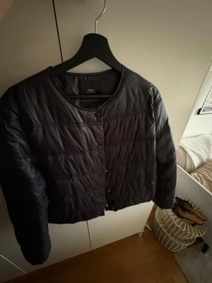 Svart kort pufferjacka från Zara - Säljer en svart, kort pufferjacka från Zara med rund halsringning och tryckknappar framtill. Jackan har en stilren design utan huva och är lätt vadderad, perfekt för kalla vinterdagar. Passar till jeans och ger en clean look.