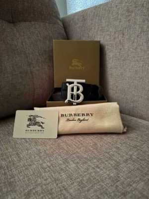 Svart Burbǝrry bälte  - Säljer ett svart bälte från Burbǝrry med en stor, silverfärgad TB-logga som spänne. Bältet kommer i originalförpackning med dustbag och äkthetskort. Perfekt accessoar för att lyfta din outfit.