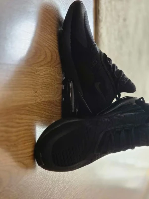 Svarta Nike Air Max 270 sneakers - Säljer ett par helsvarta Nike Air Max 270 sneakers med synlig Air-enhet i hälen och mesh-överdel för bra ventilation. Skorna har en sportig siluett, rund tå och klassisk snörning. Perfekta för dig som gillar stilrena och bekväma sneakers.